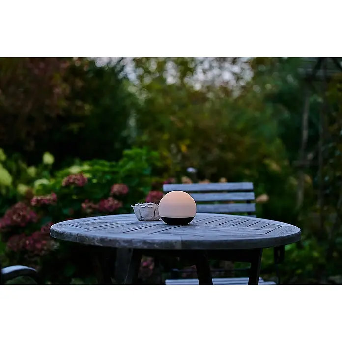 Lumix Solarleuchte Pompom - Lichtfarbe: RGB, Höhe: 100 cm, Pompom - Eine beleuchtete kugelförmige tragbare Lampe mit schwarzem Sockel auf einem hölzernen Gartentisch.