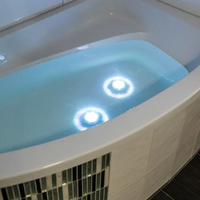LED-Poolbeleuchtung - Durchmesser: 7 cm, Transparent, LED, RGB - Weiße Eckbadewanne mit hellblauem Wasser, ausgestattet mit zwei leuchtenden Unterwasserlichtern und dekorativen Mosaikfliesen.