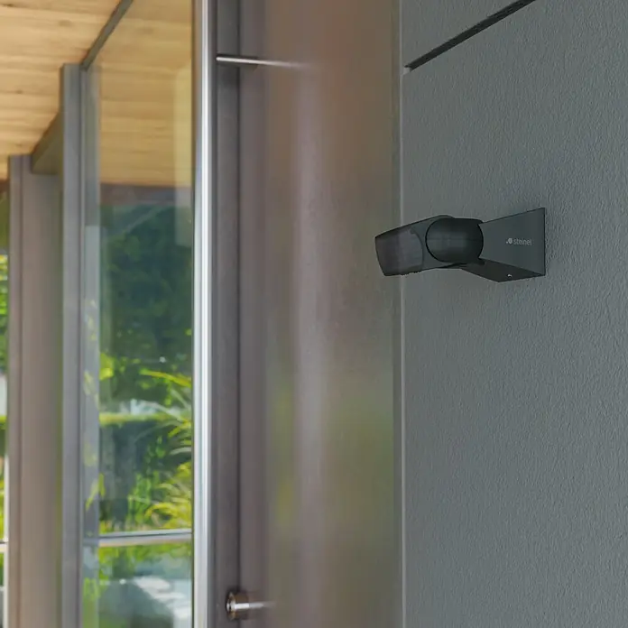 Steinel Bewegungsmelder IS 1 schwarz - Reichweite Sensor: 10 m, Schwarz, Erfassungswinkel Sensor: 120 °, L x B x H: 120 x 80 x 50 mm - Dunkelgrauer Steinel Bewegungsmelder an einer Wand neben einem modernen Gebäudeeingang, ideal für Sicherheit und Beleuchtung im Außenbereich.
