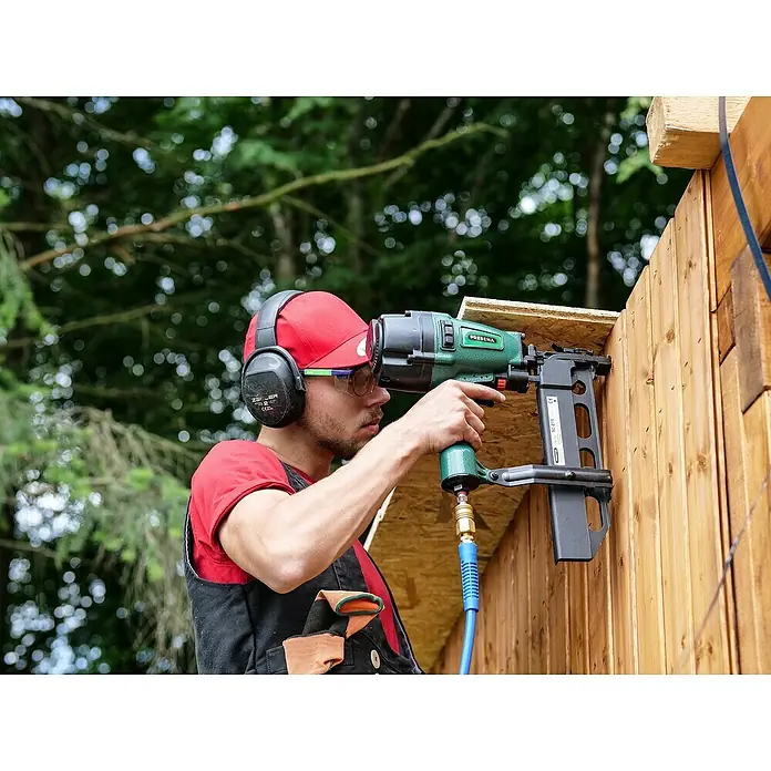 Prebena Druckluftnagler 5C-Z75 - Betriebsdruck: 5 bar - 8 bar, Passend für: Prebena Heftklammern der Type Z von 35 - 75 mm - Mann verwendet eine grüne Prebena Druckluftnagelpistole mit blauem Schlauch, um Holzbretter zu befestigen, mit Schutzausrüstung.