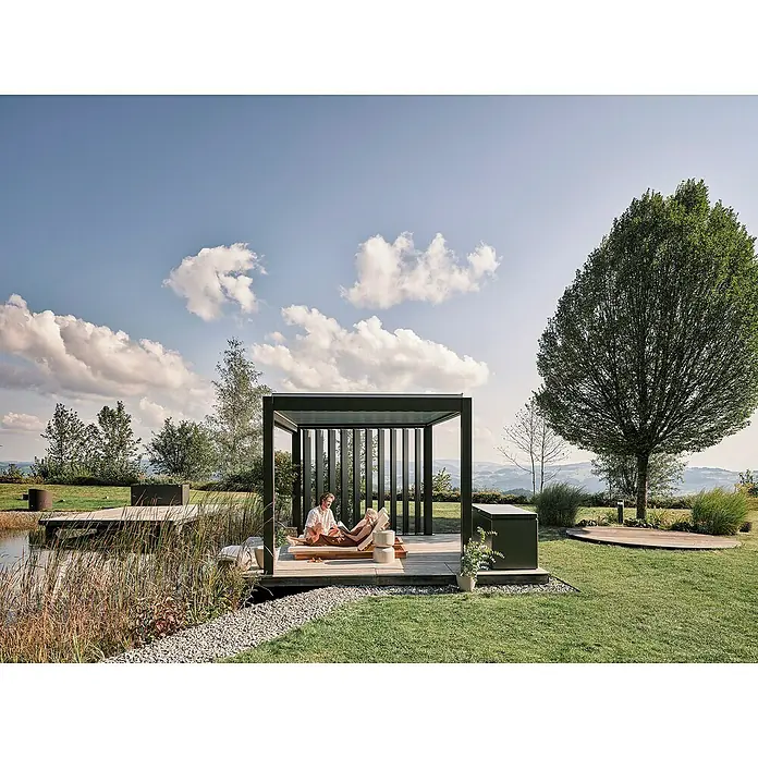 Biohort Seitenwand - Passend für Biohort Lamellenpavillon 4 m, Dunkelgrau Metallic - Eine moderne schwarze Pergola schützt zwei Personen, die auf einer Holzterrasse an einem Teich entspannen, mit Hügeln und Bäumen im Hintergrund.