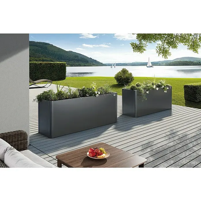 Biohort Belvedere Hochbeet - 53 x 201 x 77 cm, Stahl, Dunkelgrau Metallic - Zwei große, dunkelgraue rechteckige Pflanzkübel auf einer Holzterrasse, gefüllt mit grünen Pflanzen und weißen Blumen, mit Seeblick.