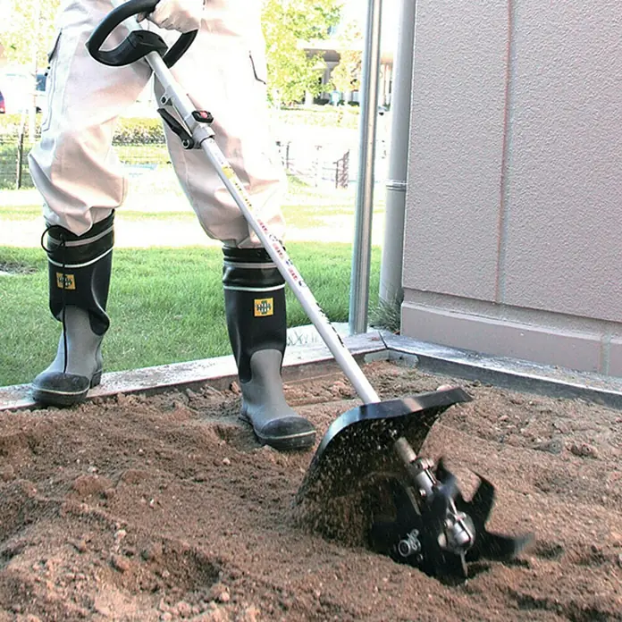 Makita Kultivator Aufsatz KR400MP - 16 cm Arbeitsbreite - Person in Arbeitsstiefeln nutzt eine Mini-Gartenhacke mit schwarzen Klingen zur Bodenbearbeitung.