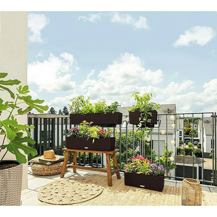 Lechuza Balconera Balkonkasten Cottage - Max. Außenlänge: 100 cm, Mokka - Dunkelbraune Pflanzgefäße in Rattanoptik auf sonnigem Balkon mit grünen Pflanzen und bunten Blumen, auf Holzbank, Boden und Geländer.