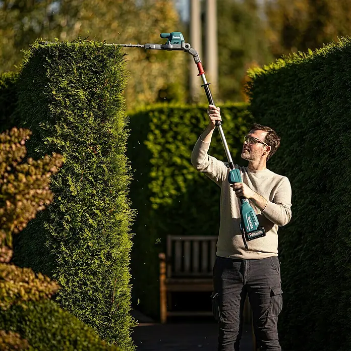 Makita XGT 40V max. Akku-Heckenschere UN001G - 40 V, Li-Ionen, Ohne Akku, Schnittlänge: 600 mm - Mann schneidet hohe Hecke mit teal-grauem Makita Langstiel-Heckenschneider.