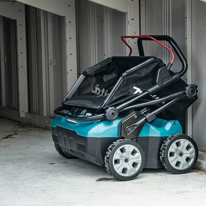 Makita XGT 40V max. Akku-Vertikutierer UV001GZ - Anzahl Akkus: Ohne Akku, 40 V, Arbeitsbreite: 38 cm - Türkis-schwarzer Rasenmäher mit schwarzem Fangkorb aus Netzstoff und roten Griffakzenten, in einem Raum mit Metallwand gelagert.