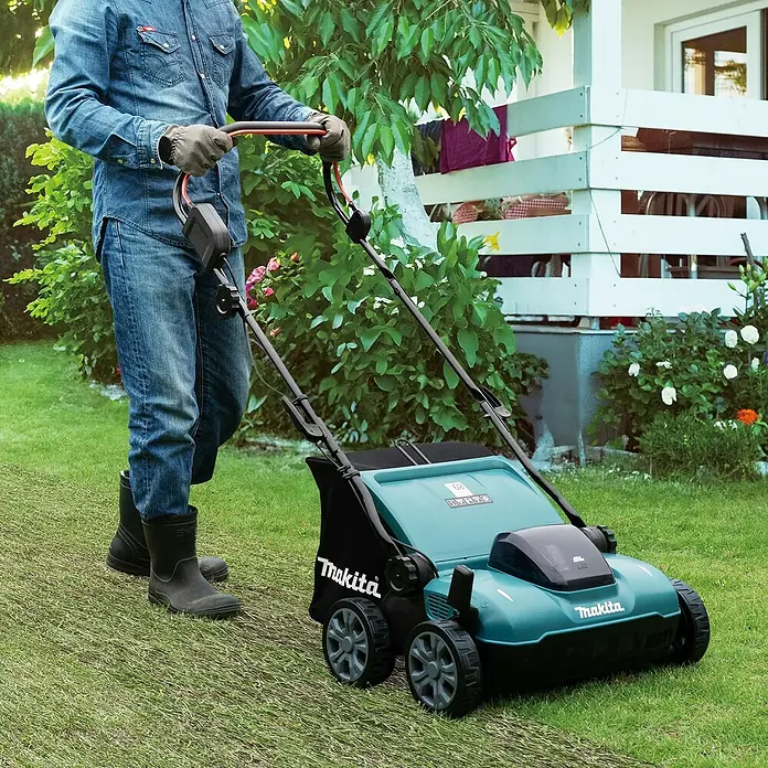 Makita LXT 18V Akku-Vertikutierer DUV320Z - Anzahl Akkus: Ohne Akku, 18 V, Arbeitsbreite: 32 cm - Eine Person in Jeans schiebt einen türkis-schwarzen Makita Rasenmäher und mäht Gras im Garten.