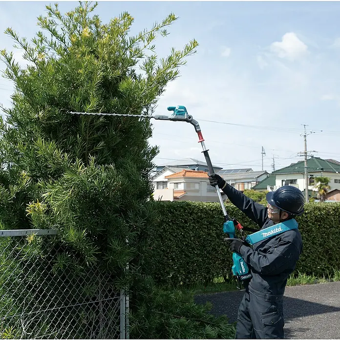 Makita XGT 40V max. Akku-Heckenschere UN001G - 40 V, Li-Ionen, Ohne Akku, Schnittlänge: 600 mm - Eine Person trimmt mit einer türkisfarbenen und grauen Makita Langstiel-Heckenschere einen hohen Busch und trägt Schutzkleidung sowie einen Gurt.