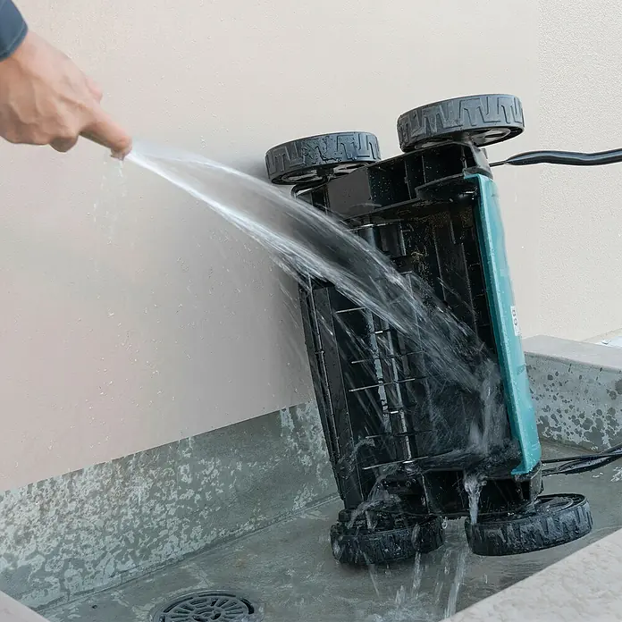 Makita LXT 18V Akku-Vertikutierer DUV320Z - Anzahl Akkus: Ohne Akku, 18 V, Arbeitsbreite: 32 cm - Person reinigt einen umgedrehten, schwarz-türkisen Rasenmäher mit einem Schlauch, Wasser spritzt auf die Unterseite.