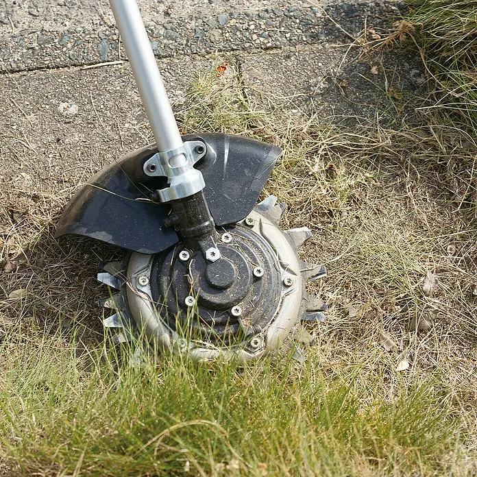 Makita Gras- & Strauchscherenaufsatz EM407MP - Passend für: Alle Makita Akku- und Benzin-Multifunktionsantriebe - Schwarzer und silberner Freischneiderkopf mit gezahnter Metallklinge, auf Gras und Beton liegend.