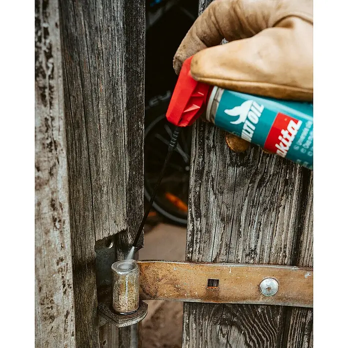 Makita Pflegespray - 0,3 l - Behandschuhte Hand sprüht Makita Multi Öl aus einer türkis-roten Dose auf ein rostiges Metalltorband.