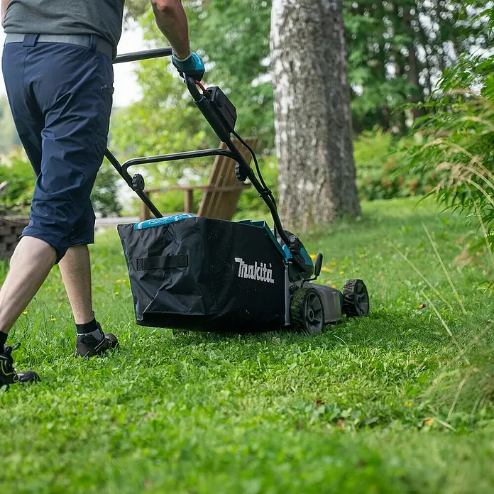 Makita LXT 18V Akku-Rasenmäher DLM466PT2 - 18 V, 2 Akkus, 5 Ah, Schnittbreite: 46 cm - Eine Person mäht mit einem Makita Rasenmäher mit schwarzem Fangkorb grünes Gras im Garten.