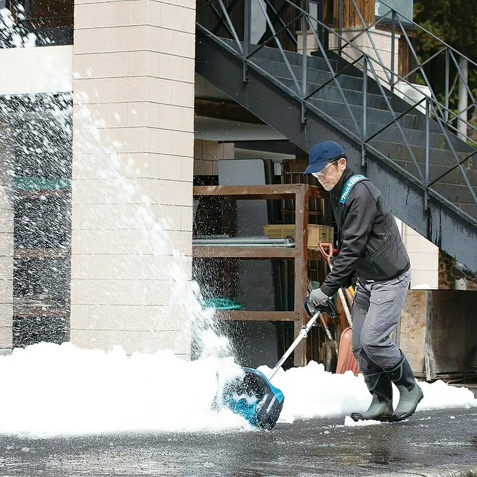 Makita Multifunktionswerkzeug-Zubehör Schneefräsen-Aufsatz SN400MP - Passend für: DUX18, DUX60, UX01G - Person räumt mit einer blau-schwarzen Schneeschaufel Schnee von einem gepflasterten Bereich; Schnee fliegt von der Schaufel.