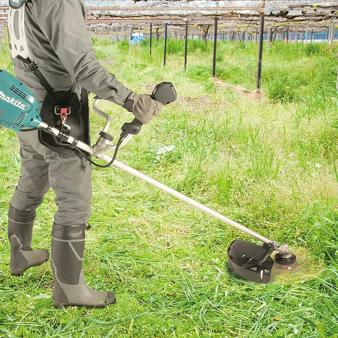 Makita XGT 40V max. Akku-Freischneider UR012GZ06 - 40 V, 8 Ah, Ohne Akku, Schnittbreite Faden: 305 cm - Person bedient einen türkis-silbernen Makita Rasentrimmer zum Grasschneiden auf einem Feld, trägt Gurtzeug und Stiefel.
