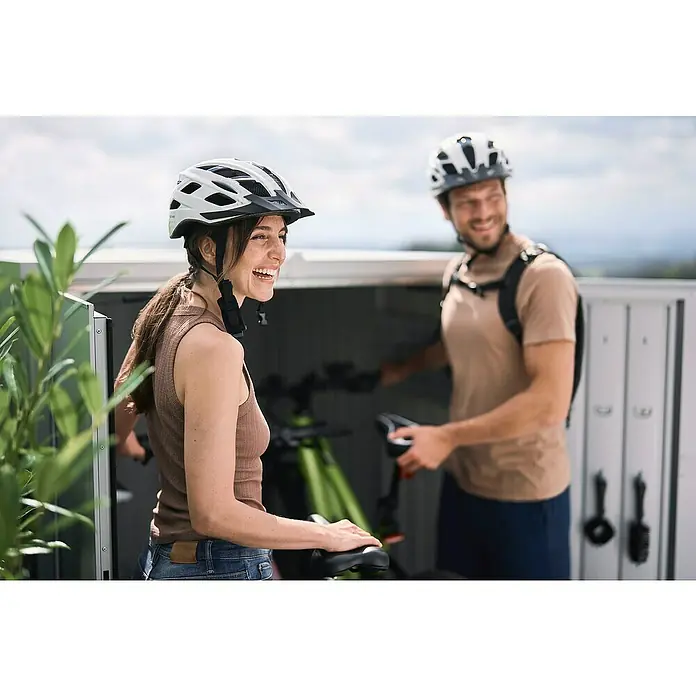 Biohort MiniGarage Fahrradgarage L - Dunkelgrau Metallic, Außenbreite inkl. Dachüberstand: 145 cm, Außentiefe inkl. Dachüberstand: 203 cm - Zwei lächelnde Radfahrer mit weißen Helmen holen ihre grünen Fahrräder aus dem Abstellraum.