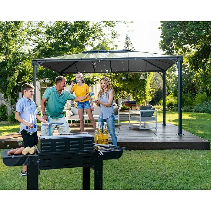 Palram – Canopia Pavillon Martinique 4300 - B x T: 430 x 295 cm, Farbe Gestell: Anthrazit, Aluminium - Dunkelgrauer Pavillon mit transparenten Dachplatten spendet Schatten für ein Familienfest auf einer sonnigen Terrasse.
