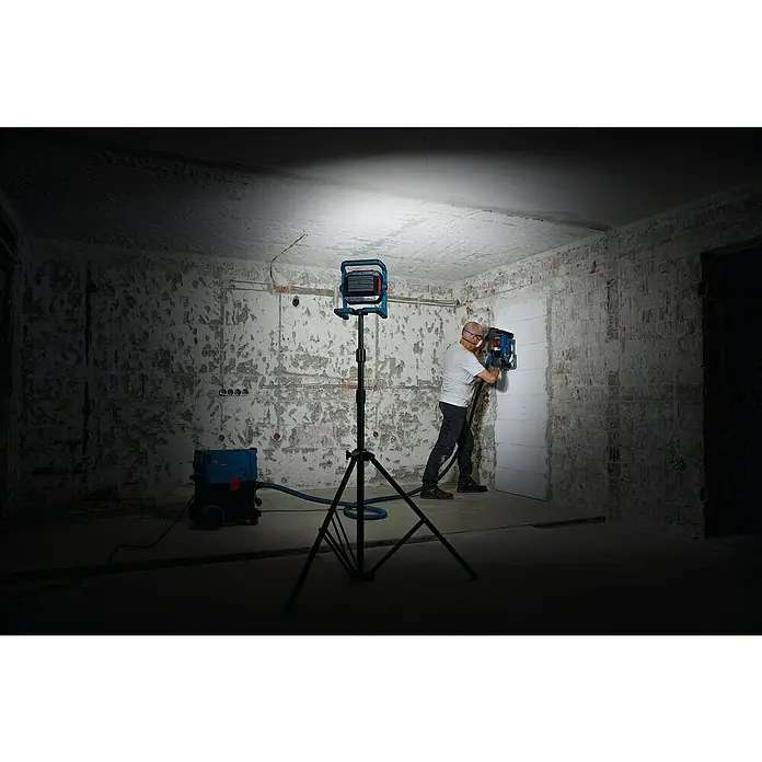 Bosch Professional AMPShare 18V LED-Akkustrahler GLI 18V-5000 C - 18 V, Ohne Akku, Lichtstrom: 5.000 lm - Arbeiter bearbeitet Wand mit blauem Elektrowerkzeug, verbunden mit Staubabsauger, beleuchtet von Bosch-Arbeitsleuchte auf Stativ.