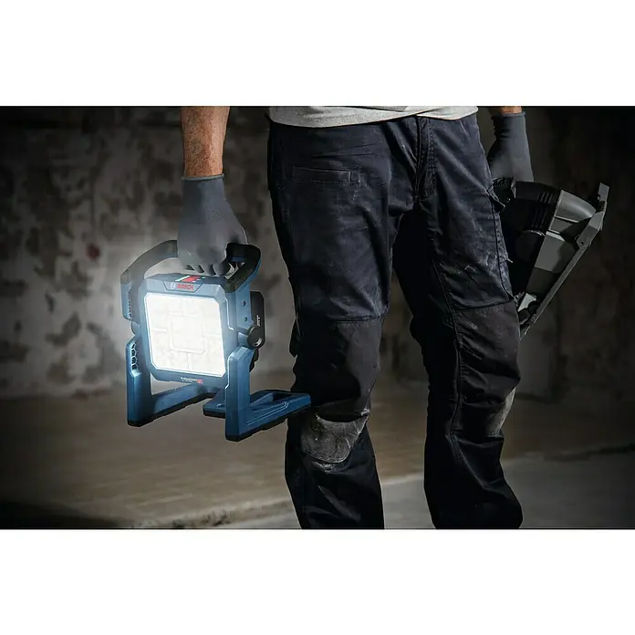 Bosch Professional AMPShare 18V LED-Akkustrahler GLI 18V-5000 C - 18 V, Ohne Akku, Lichtstrom: 5.000 lm - Eine Person in Arbeitshandschuhen hält eine leuchtende blaue Bosch Arbeitsleuchte in einem dunklen Arbeitsbereich.