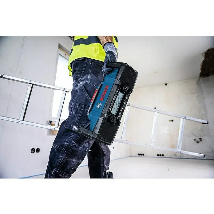 Bosch Professional AMPShare 18V Akku-Radio GPB 18V- 3 SC - 18 V, Ohne Akku - Ein Arbeiter trägt ein blau-schwarzes Bosch Baustellenradio mit Digitalanzeige neben einer Leiter auf einer Baustelle.