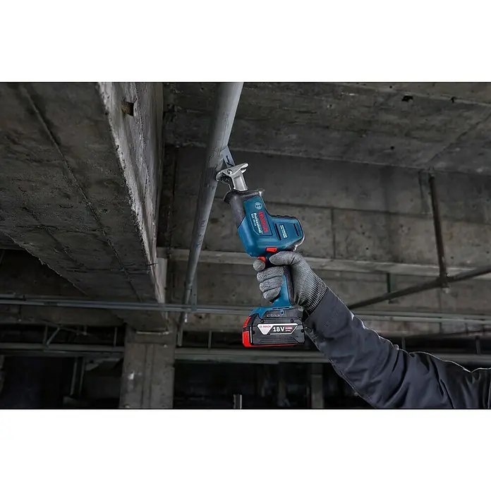 Bosch Professional AMPShare 18V Akku-Säbelsäge GSA 18V-24 -  - Behandschuhte Hand hält blaue und schwarze Bosch 18V Säbelsäge mit 4,0Ah Akku, die ein Metallrohr vor Betonhintergrund schneidet.