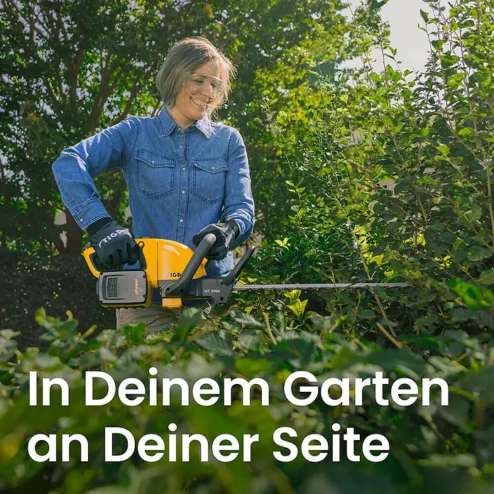 Stiga Akku-Heckenschere HT 300E - 2 Akkus, Schnittlänge: 54 cm - Frau in Jeanshemd und Schutzbrille schneidet grüne Hecken mit einer gelb-schwarzen STIGA Heckenschere im Garten.