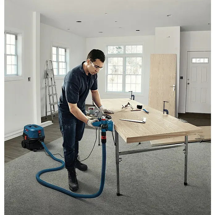 Bosch Professional Elektrohobel GHO 20-82 - 700 W, Hobeltiefe: 0 mm - 2 mm, Leerlaufdrehzahl: 16.500 U/min - Mann benutzt blau-schwarzes Elektrowerkzeug mit Staubabsaugung auf Holz, verbunden mit blauem Schlauch zu blau-schwarzem Industriesauger.
