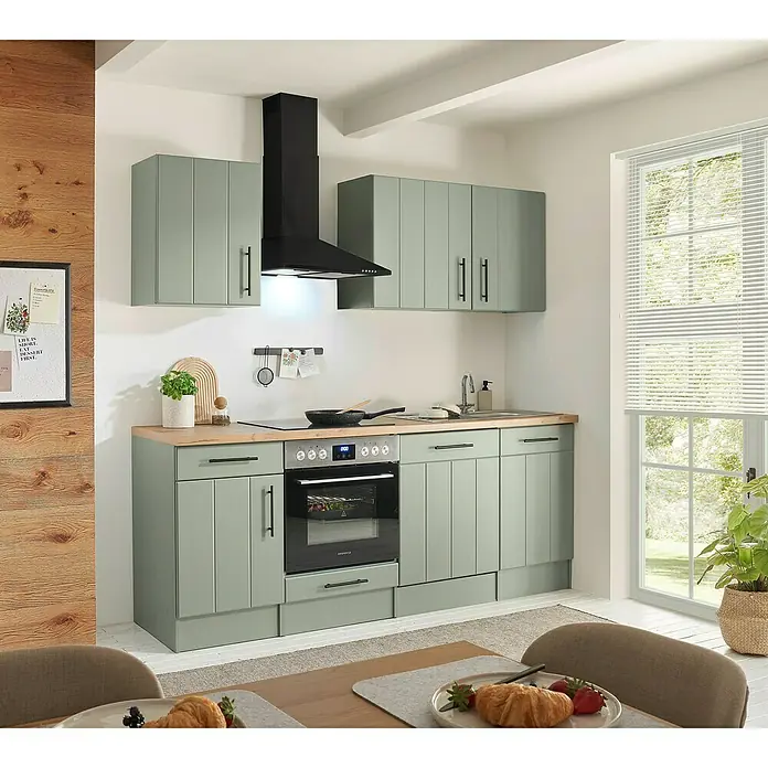 Respekta Küchenzeile Anton Typ 1 - Breite: 220 cm, Schilfgrün, Mit Elektrogeräten - Moderne Küche mit salbeigrünen Unter- und Oberschränken, heller Holz-Arbeitsplatte, schwarzem Backofen, Kochfeld und Dunstabzugshaube. Esstisch im Vordergrund.