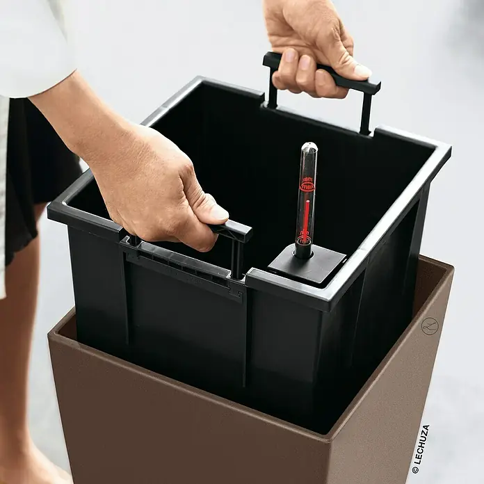 Lechuza Color Pflanzsäule eckig Cubico - Außenmaß (L x B x H): 30 x 30 x 56 cm, Kunststoff, Muskat - Zwei Hände setzen einen schwarzen rechteckigen Pflanzeinsatz mit rotem Wasserstandsanzeiger in einen braunen Pflanzkübel ein.