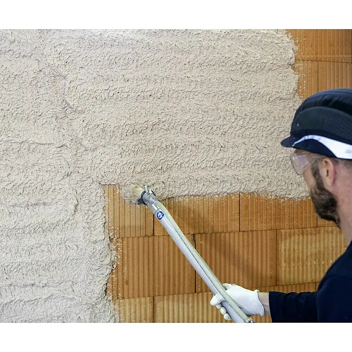 Knauf Maschinenputz MP 75 L - 30 kg, Innen - Ein Bauarbeiter verwendet eine Spritzpistole, um helles, strukturiertes Dämmmaterial auf eine Ziegelmauer aufzutragen.