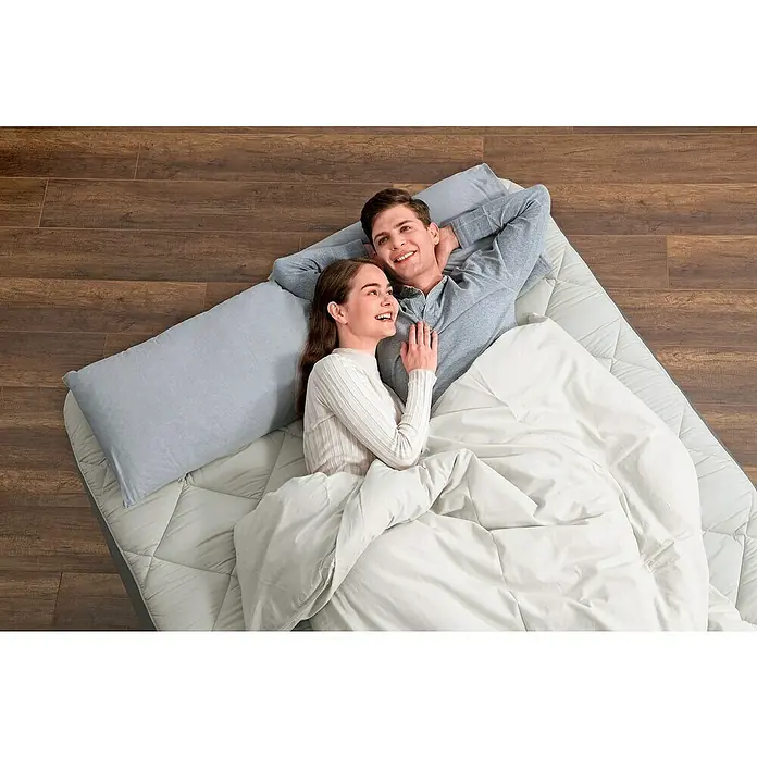 Bestway Luftbett - L x B x H: 203 x 152 x 51 cm, Dunkelbraun, Integrierte Luftpumpe - Lächelndes Paar ruht auf gestepptem Matratzentopper und weißer Bettdecke auf Holzboden.