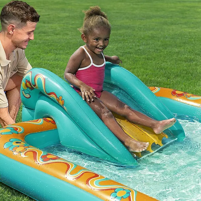 Bestway Planschbecken Retro-Welt - 152 x 193 x 74 cm - Kind rutscht eine farbige, türkis-orange aufblasbare Wasserrutsche in ein Planschbecken hinunter, ein Erwachsener schaut zu.