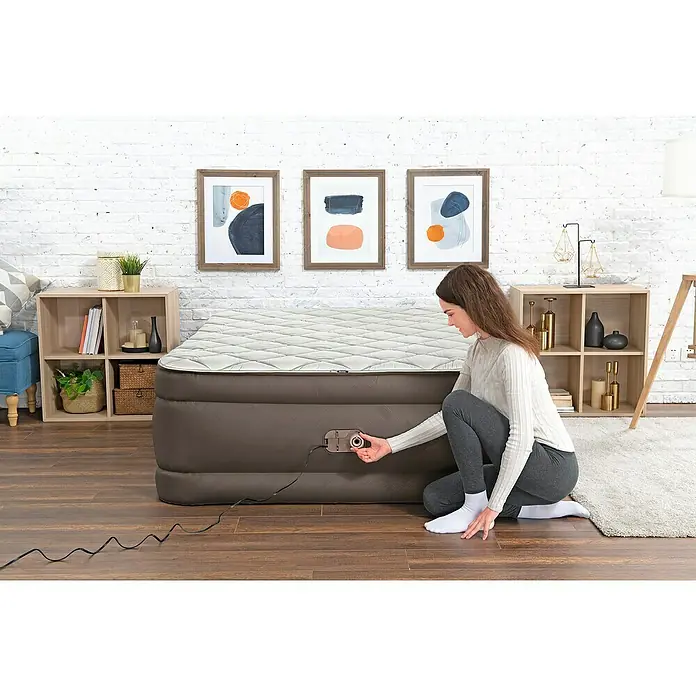 Bestway Luftbett - L x B x H: 203 x 152 x 51 cm, Dunkelbraun, Integrierte Luftpumpe - Frau bedient die integrierte Pumpe einer großen grauen Luftmatratze mit weißer Steppoberfläche. Sie kniet daneben und stellt das Rad ein.