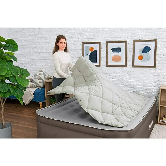 Bestway Luftbett - L x B x H: 203 x 152 x 51 cm, Dunkelbraun, Integrierte Luftpumpe - Frau legt einen hellen, gesteppten Topper auf eine graue Luftmatratze.