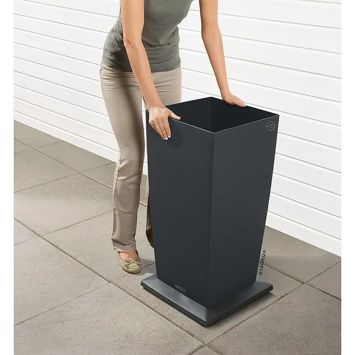 Lechuza Pflanzenroller Quadratisch - L x B x H: 41 x 41 x 4,5 cm, Kunststoff, Anthrazit Metallic - Frau platziert einen dunkelgrauen rechteckigen Pflanzkübel auf den passenden Sockel.