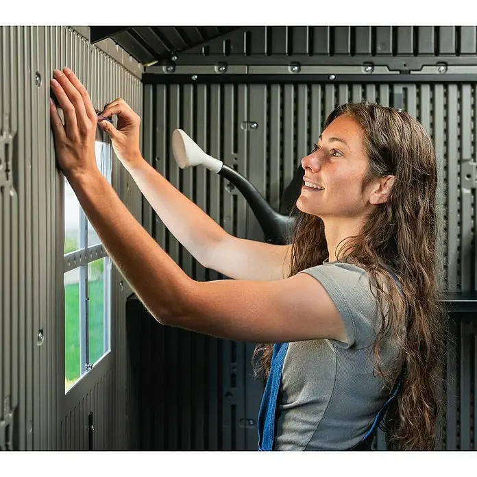 Lifetime Gerätehaus Kentucky - Kunststoff, Grau, Außenmaß inkl. Dachüberstand (B x T): 427 x 206 cm - Frau montiert ein transparentes Fenster in die graue geriffelte Kunststoffwand eines Schuppens.