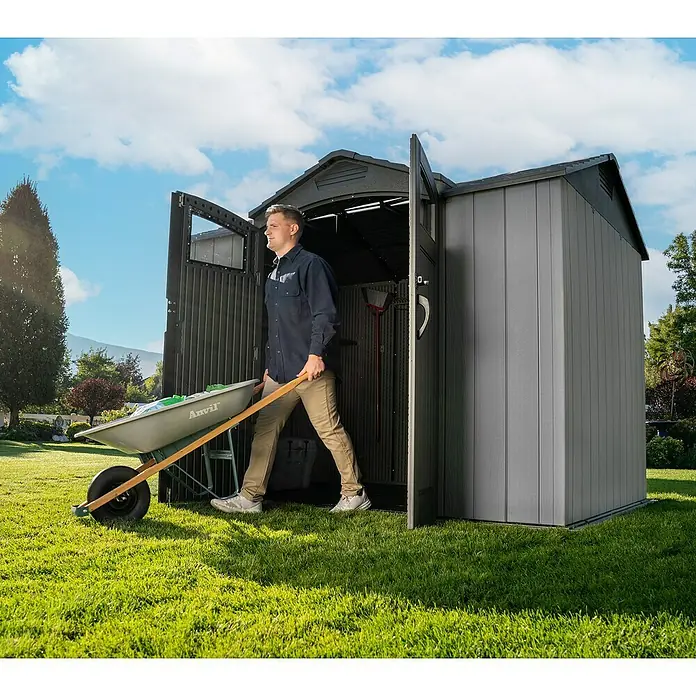 Lifetime Gerätehaus Ohio - Kunststoff, Grau, Außenmaß inkl. Dachüberstand (B x T): 283,5 x 214,1 cm - Dunkelgraues Gartenschuppen aus Kunststoff, ein Mann fährt mit einer Schubkarre heraus, zeigt geräumige und zugängliche Lagerung.