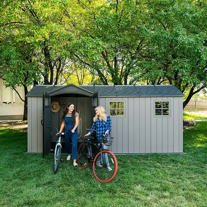 Lifetime Gerätehaus Idaho - Kunststoff, Grau, Außenmaß inkl. Dachüberstand (B x T): 498 x 214 cm - Zwei Frauen verstauen Fahrräder in einem grauen, robusten Gartenschuppen. Der Schuppen ist wetterfest und funktional.