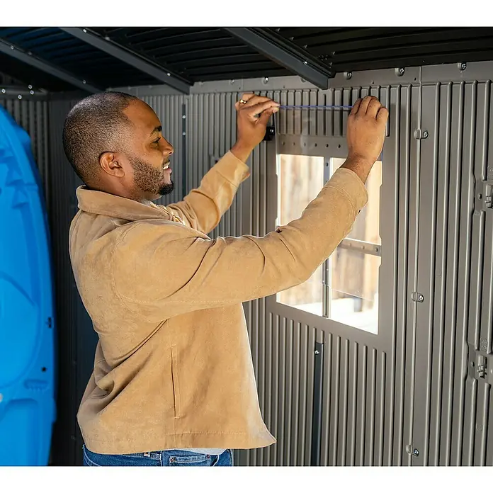 Lifetime Gerätehaus Utah - Kunststoff, Grau, Außenmaß inkl. Dachüberstand (B x T): 206 x 427 cm - Mann installiert Zubehör in einem grauen Kunststoff-Gartenschuppen.
