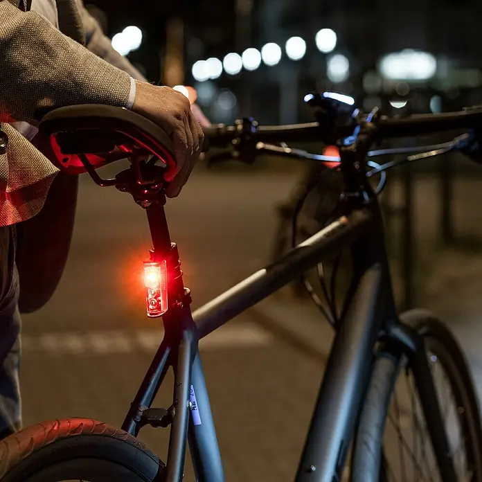 Sigma Dynamo-LED-Rücklicht Blaze - Ohne Batterien - Eine Person steht nachts neben einem Fahrrad, dessen hellrotes Rücklicht für Sichtbarkeit leuchtet.