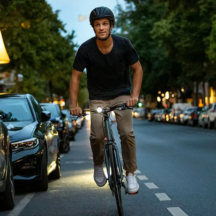 Sigma Fahrradbeleuchtungs-Set Aura 30/Curve - 2 -tlg., Mit Batterien - Mann fährt nachts Fahrrad, trägt einen schwarzen Helm, mit hellem Frontlicht, das die Straße beleuchtet.