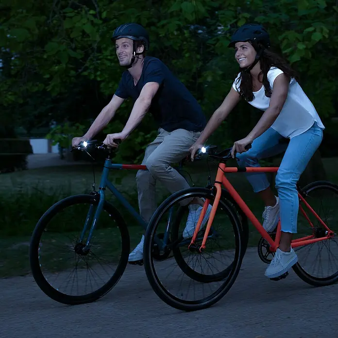 Sigma Fahrradbeleuchtungs-Set Aura 30/Curve - 2 -tlg., Mit Batterien - Zwei Personen mit Helmen fahren nachts Fahrräder mit hellen Frontlichtern.