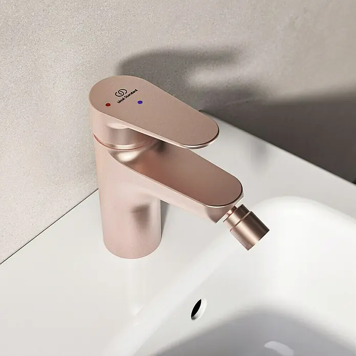 Ideal Standard Alu+ Bidetarmatur - Höhe: 140 mm, Rosé - Roségoldener Ideal Standard Bidet-Mischbatterie mit Heiß- und Kaltwasseranzeigen, auf einem weißen Bidetbecken montiert.