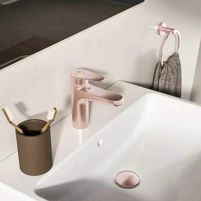Ideal Standard Alu+ Waschtischarmatur - Höhe: 160 cm, Rosé - Roségoldener Badarmatur mit passendem Waschbeckenablauf und Handtuchring, neben einem weißen Keramikwaschbecken.