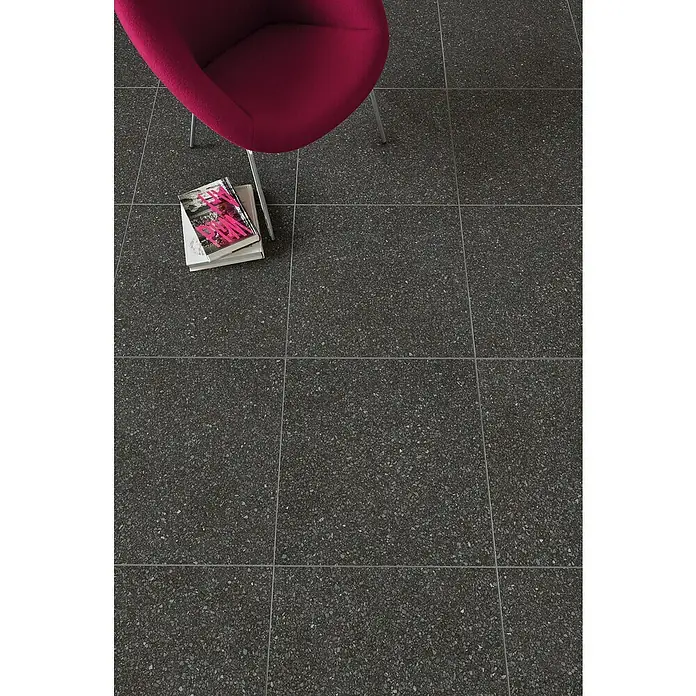 DryTile Feinsteinzeugfliese Terrazzo Black - 60 x 60 cm, Schwarz, Matt - Dunkelgraue, gesprenkelte Bodenfliesen im Quadrat mit heller Fuge, unter magenta Stuhl und Büchern.