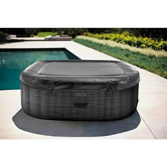 Intex Pure Spa Mobil-Whirlpool Greystone Deluxe - L x B x H: 196 x 196 x 71 cm, Grau, 1.098 l - Dunkelgrauer aufblasbarer rechteckiger Whirlpool mit gemusterter Außenfläche und passender Abdeckung, draußen auf einer Terrasse neben einem Pool.