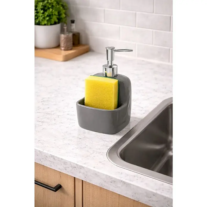Versa Dispensador de jabón Aqua Karslen - Cerámica Soft-Touch, Gris - Dispensador de jabón gris con bomba cromada y soporte integrado para una esponja amarilla brillante, junto a un fregadero de acero en una encimera de cocina.