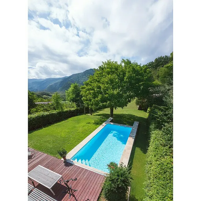 Mountfield Bausatz-Pool - L x B x H: 490 x 350 x 140 cm, 17,59 m³, Weiß - Ein rechteckiger blauer Swimmingpool in einem üppigen grünen Garten mit Holzterrasse und fernen Bergen unter bewölktem Himmel.
