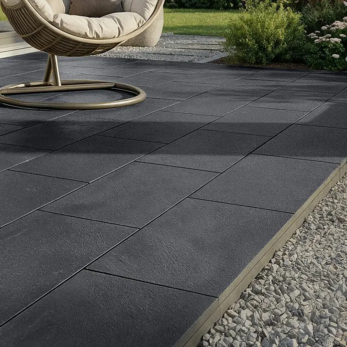 EHL Terrassenplatte Mesafino - 80 x 40 x 4 cm, Basalt, Beton - Dunkelgraue, texturierte Terrassenplatten bilden eine Außenfläche neben einem Kiesbeet.