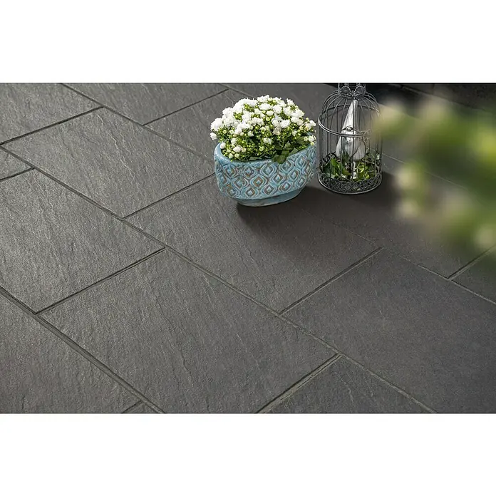 Diephaus Terrassenplatte Rancho - 60 x 30 x 4 cm, Schwarz-Basalt, Beton - Dunkelgraue, strukturierte, rechteckige Outdoor-Fliesen, die eine gepflasterte Fläche bilden, mit Topfpflanze und Dekokäfig.