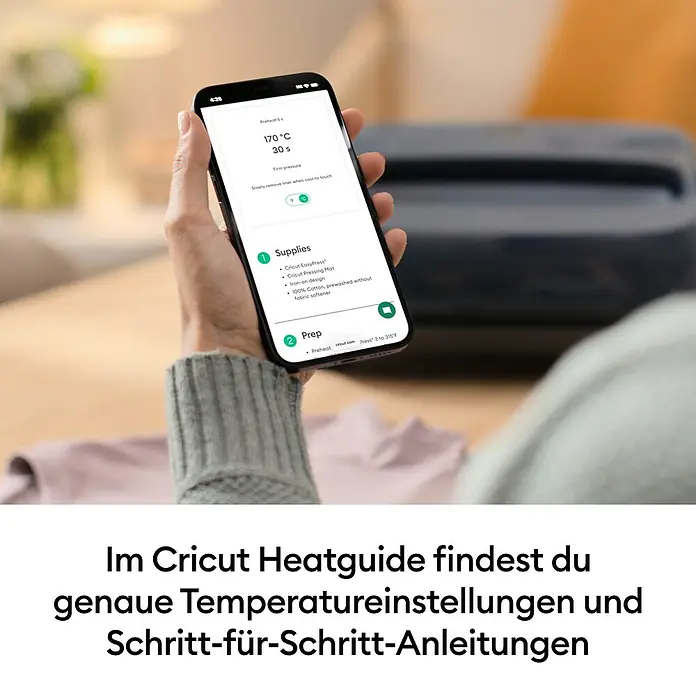 Cricut Transferpresse EasyPress SE + Hitzeschutzmatte - 25 x 30 cm, Passend für: Bügelfolien - Eine Hand hält ein Smartphone mit der Cricut Heatguide App, die genaue Temperatur- und Schritt-für-Schritt-Anleitungen zeigt, mit einer Cricut EasyPress im Hintergrund.